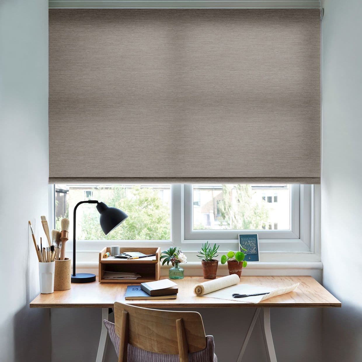 Beige textured roller blind lowered nearly fully, filtering daylight; a wooden desk beneath holds a black desk lamp, art brushes, notebooks, small potted plants, and a suburban window view.