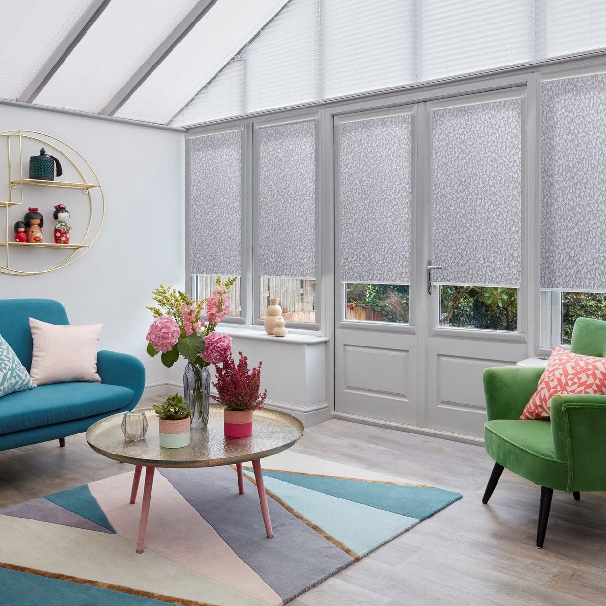 Patterned grey roller blinds cover tall conservatory windows and door, partially lowered and diffusing daylight; sunroom with pastel sofa and armchair, round coffee table, floral arrangements, geometric rug.