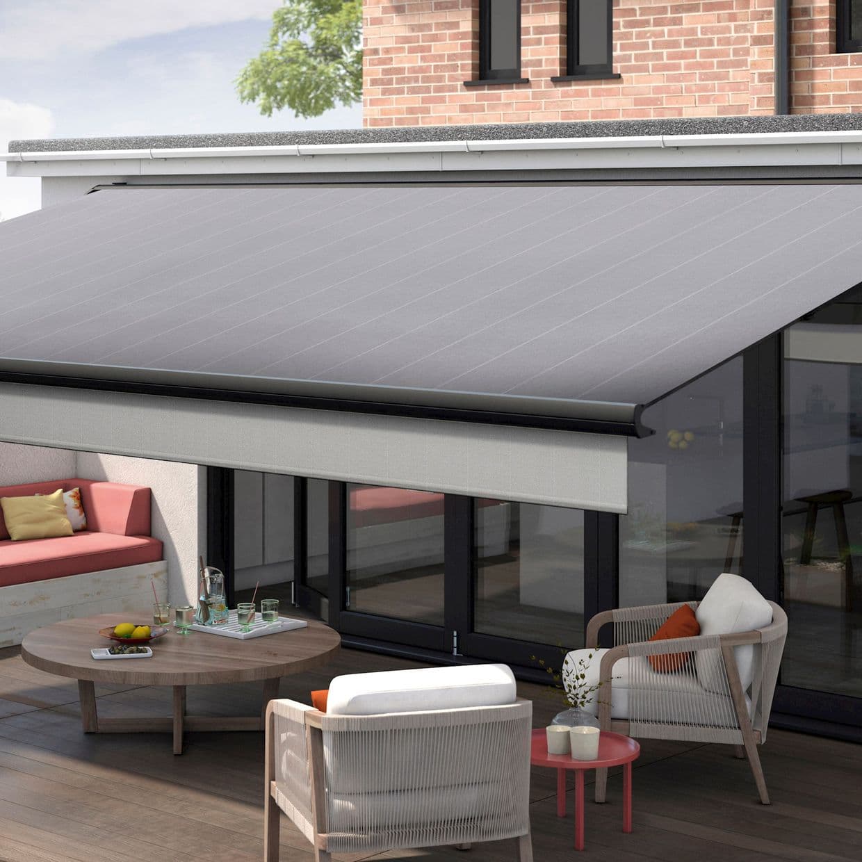 Large gray retractable awning with straight valance, fully extended over sliding glass doors, shading a modern outdoor patio with wooden deck, table, cushioned seating and brick facade in daylight.
