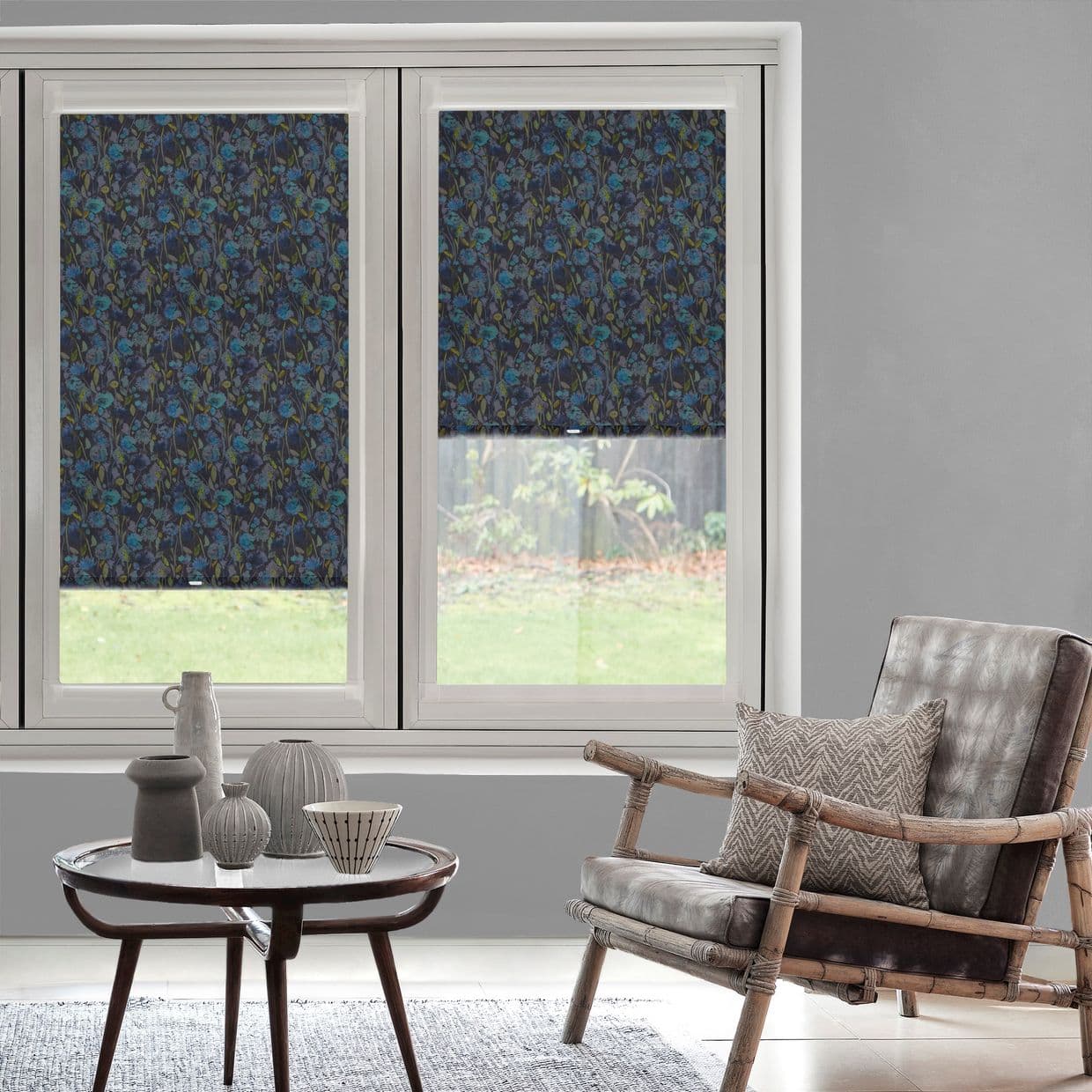 Patterned dark-blue floral roller blinds, lowered on two adjacent windows with small clear lower panels, filtering daylight; modern living room with gray walls, rattan armchair and coffee table, garden view.