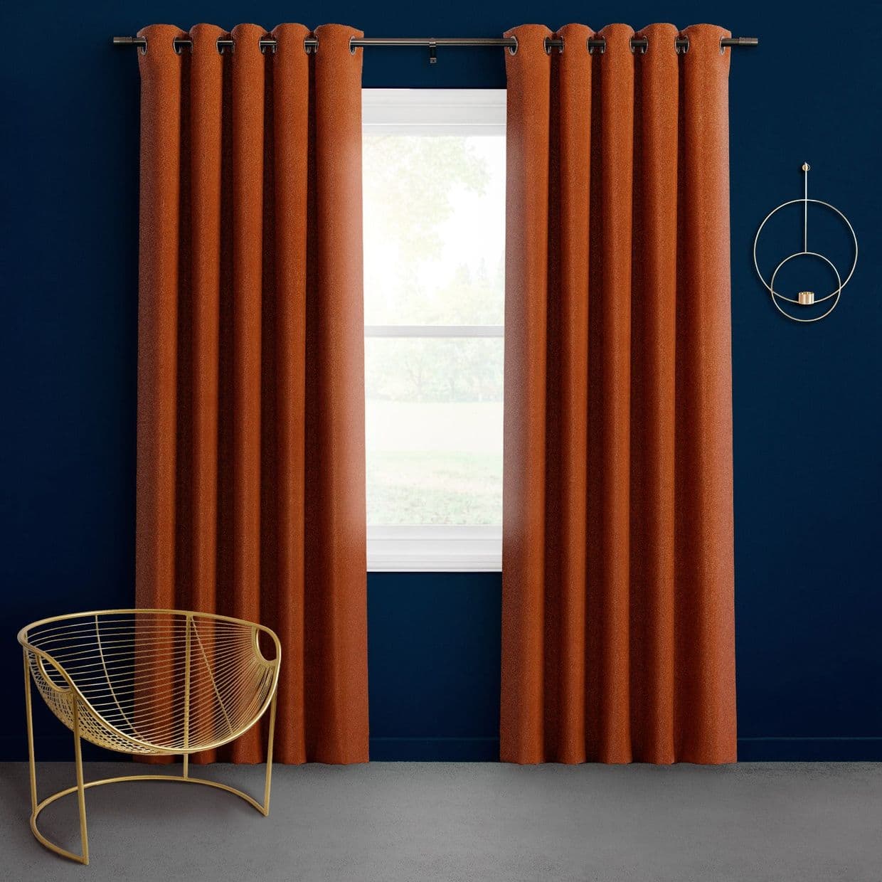 Floor-length rust-orange grommet curtains, hung on a black rod, parted slightly to reveal a bright window and diffuse daylight; navy-blue wall, gray floor, gold wire chair and circular wall sconce.