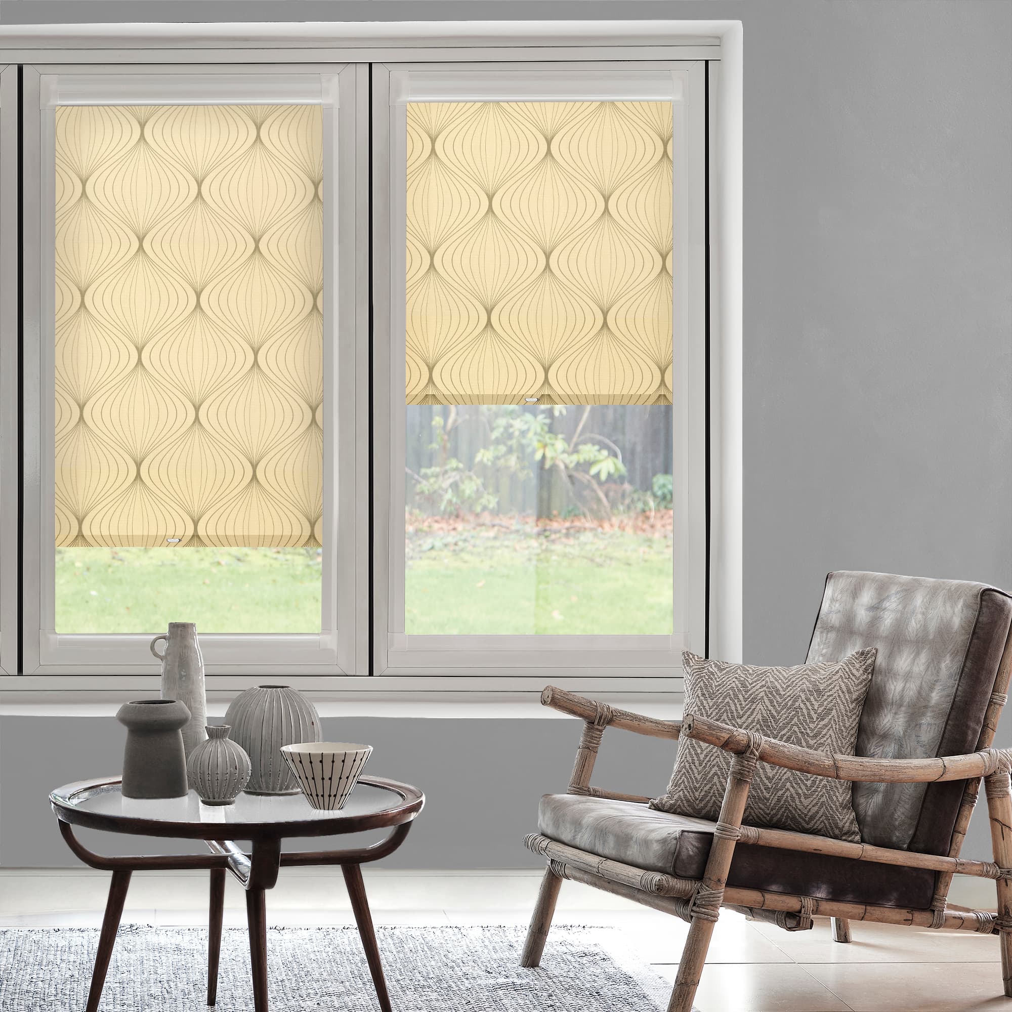 Beige roller blinds with curved pattern â€” left lowered, right partially lowered, filtering daylight into a gray living room with rattan armchair, cushion and round table with vases, grassy backyard view.