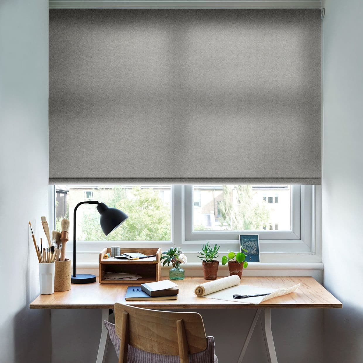 Grey textured roller blind lowered over the window, diffusing light; a small wooden desk beneath holds a lamp, brushes, notebooks, potted succulents and rolled fabric in a bright suburban room.