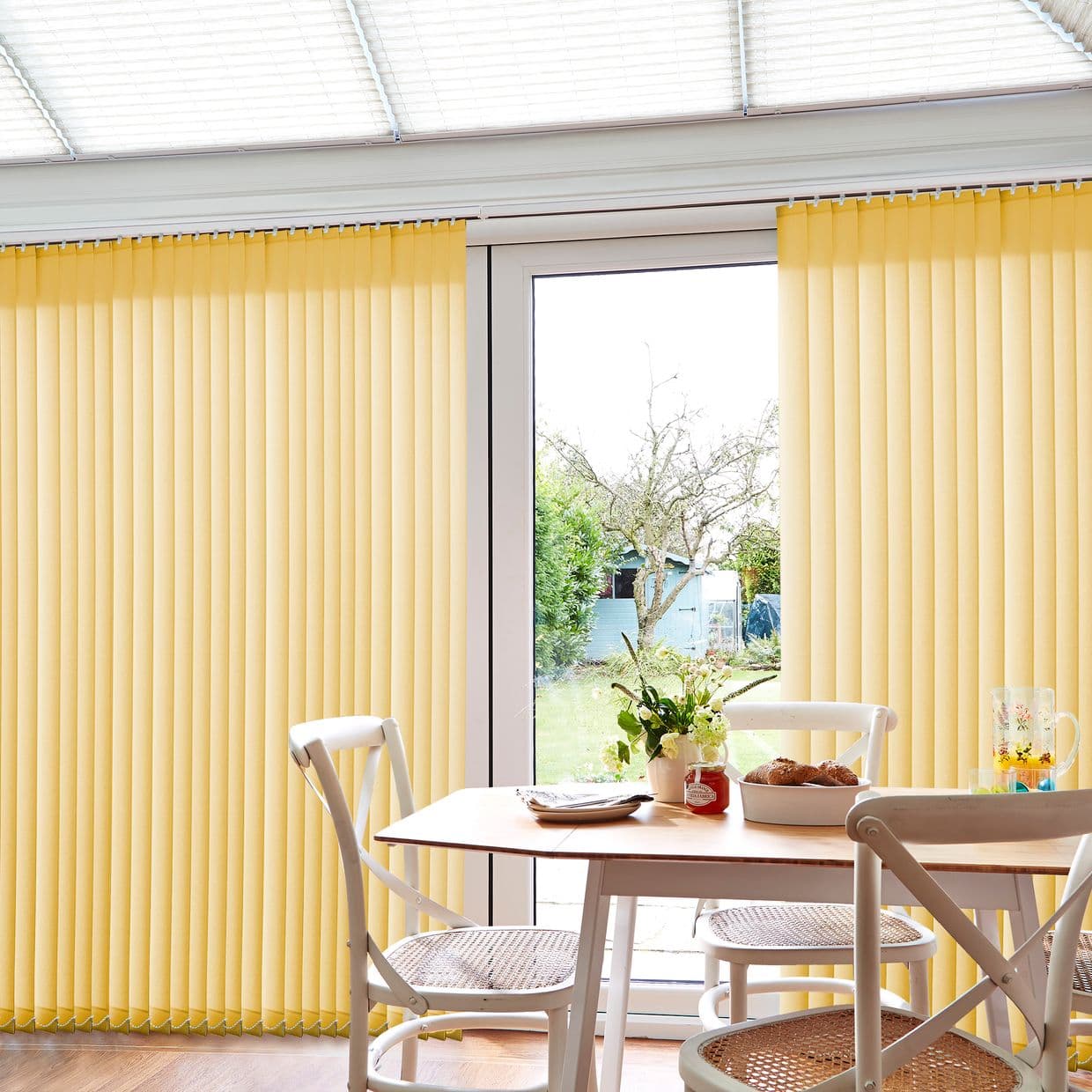 Yellow vertical blinds, pleated slats parted at the center revealing a glass patio door, filtering soft daylight into a bright dining nook with wooden table, chairs, vase, and backyard view.