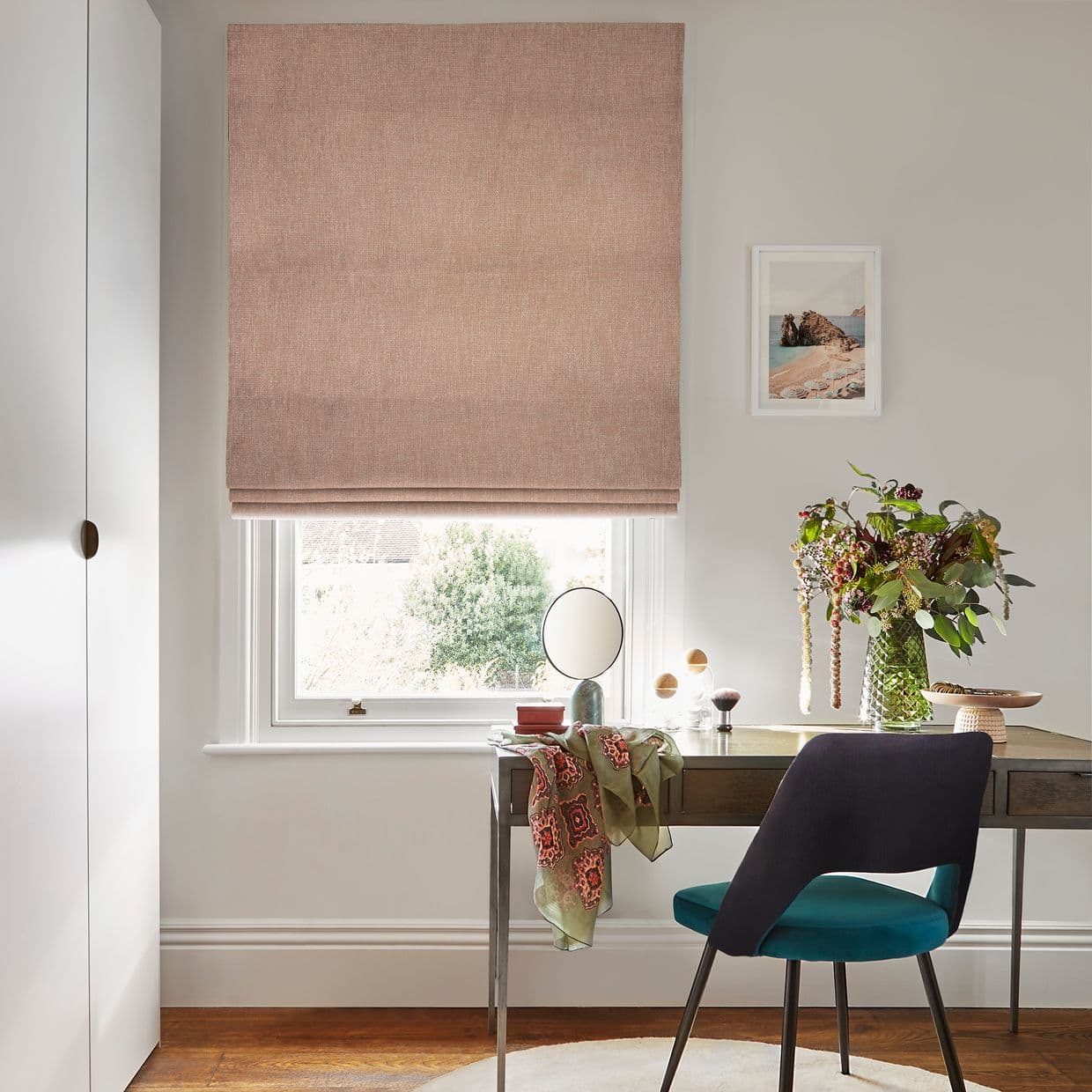 Dusty-pink Roman blind, lowered and folded across the window, filtering daylight; desk with mirror, vase of foliage, draped scarf and teal chair in a bright home office.