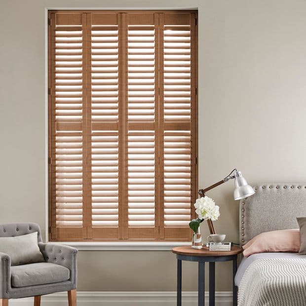 Wooden four-panel plantation shutters with horizontal louvers, closed and slightly angled to filter daylight, covering a tall window in a neutral bedroom cornerâ€”bed, bedside table, lamp and armchair.