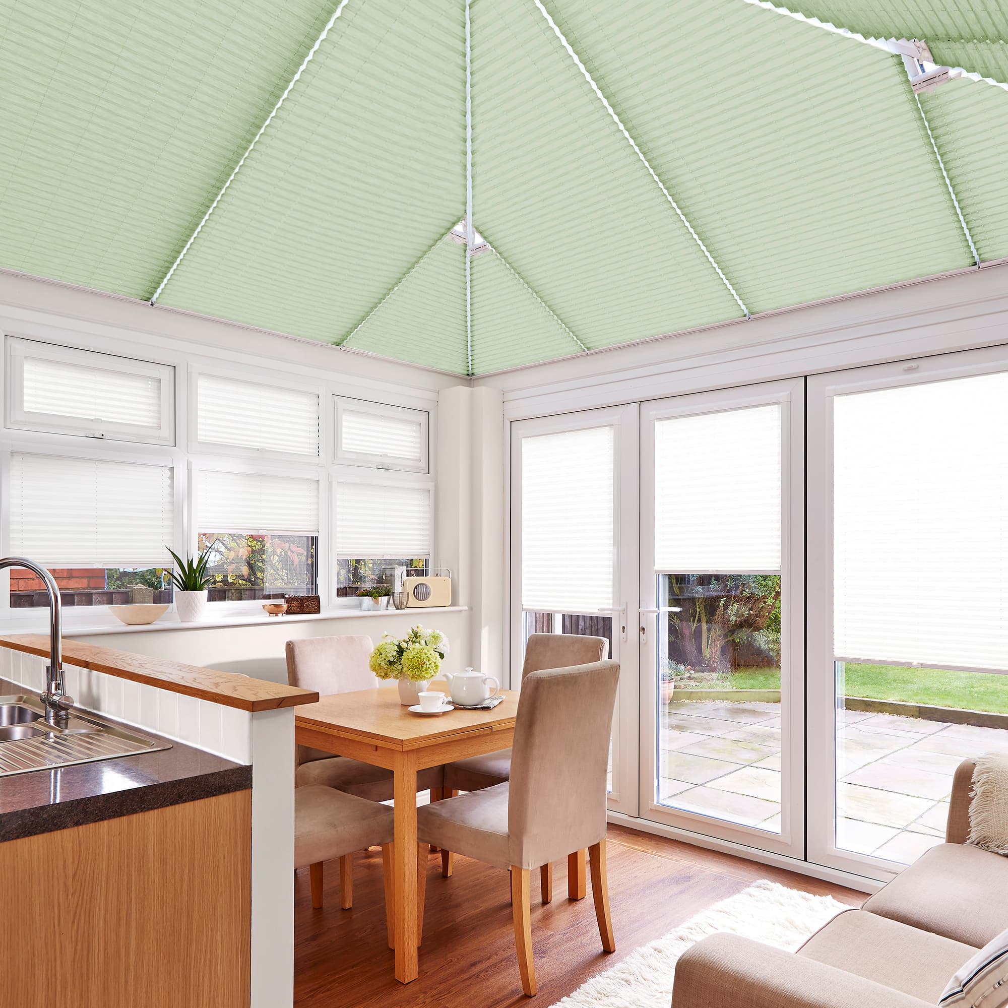Green cellular pleated roof blinds and white pleated window/door blinds, lowered and filtering daylight, in bright conservatory dining-kitchen with wooden table, upholstered chairs, sink and glass doors to patio.
