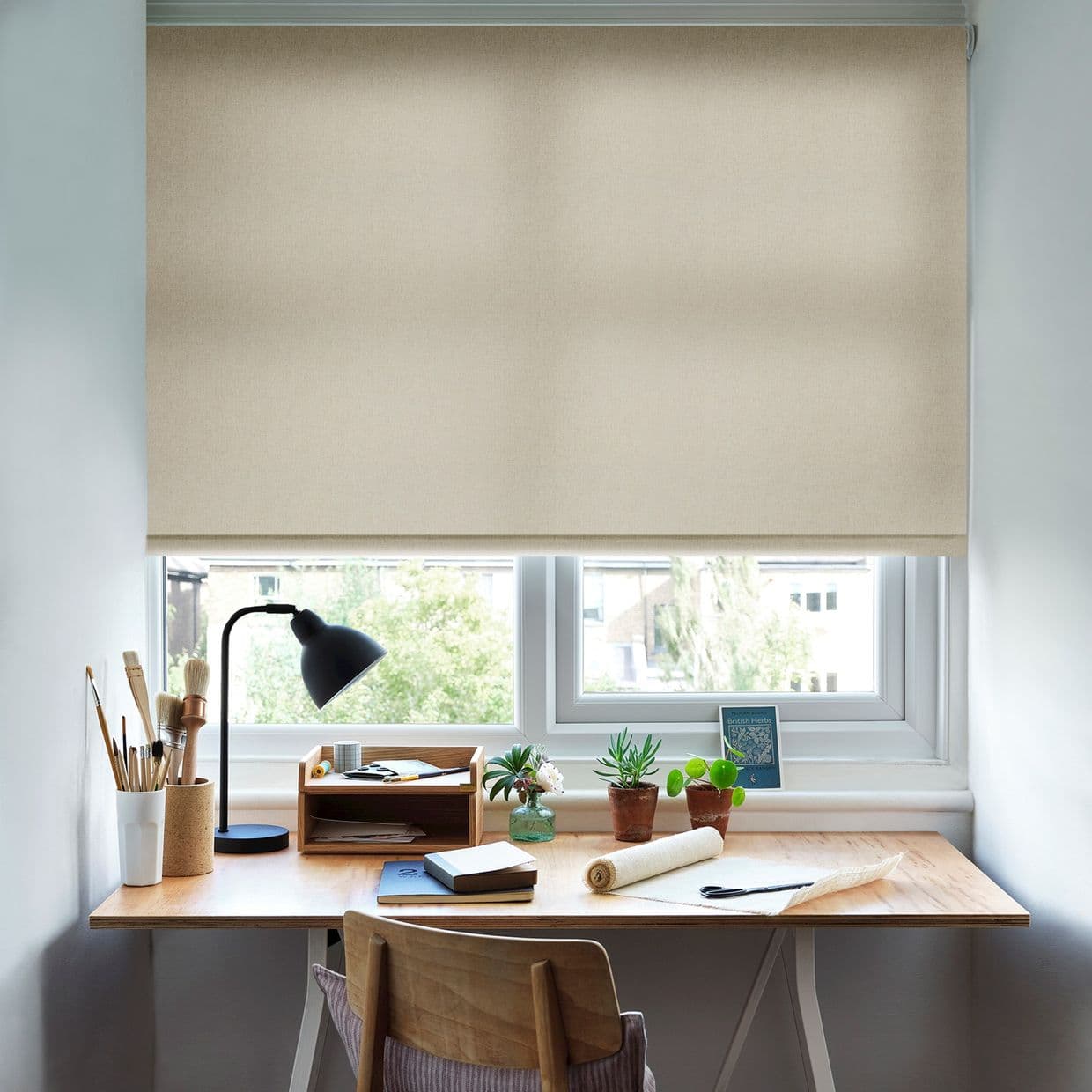 Beige fabric roller blind lowered over the window, filtering daylight and blocking the view. Below, a wooden desk workspace with lamp, art brushes, notebooks and small potted plants.