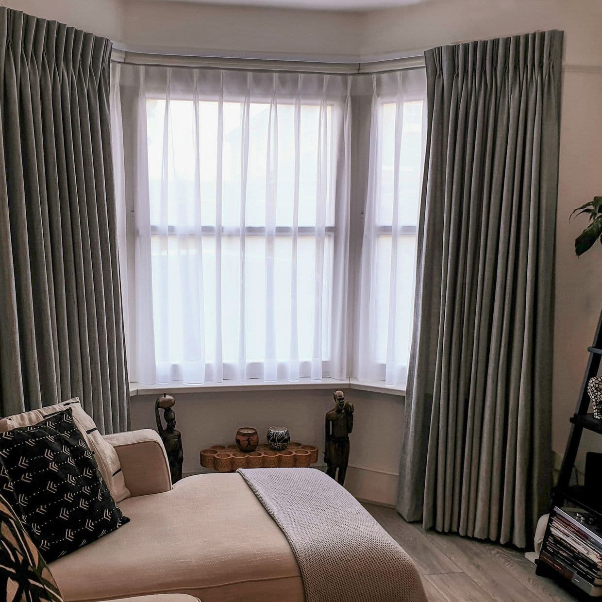Floor-length pleated grey drapes hung at either side, with translucent white sheers stretched across the bay window, filtering soft daylight into a neutral-toned living room with a chaise lounge.
