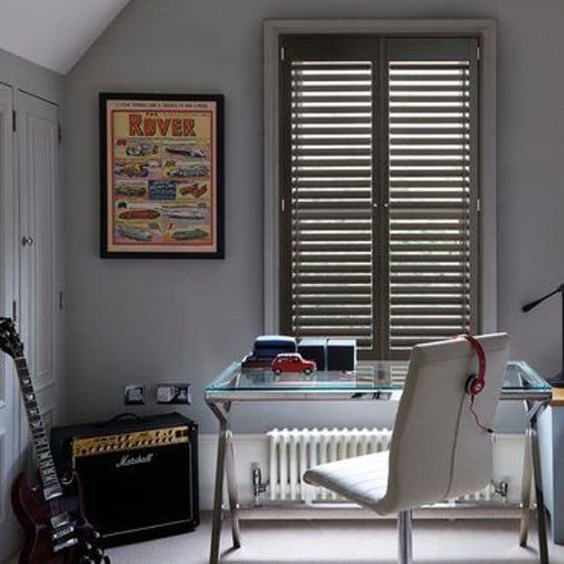 Wooden interior plantation shutters cover a double window, closed with slats angled to filter daylight; set above a glass desk in a grey home office with a chair, guitar and amplifier.

ROVER
Marshall