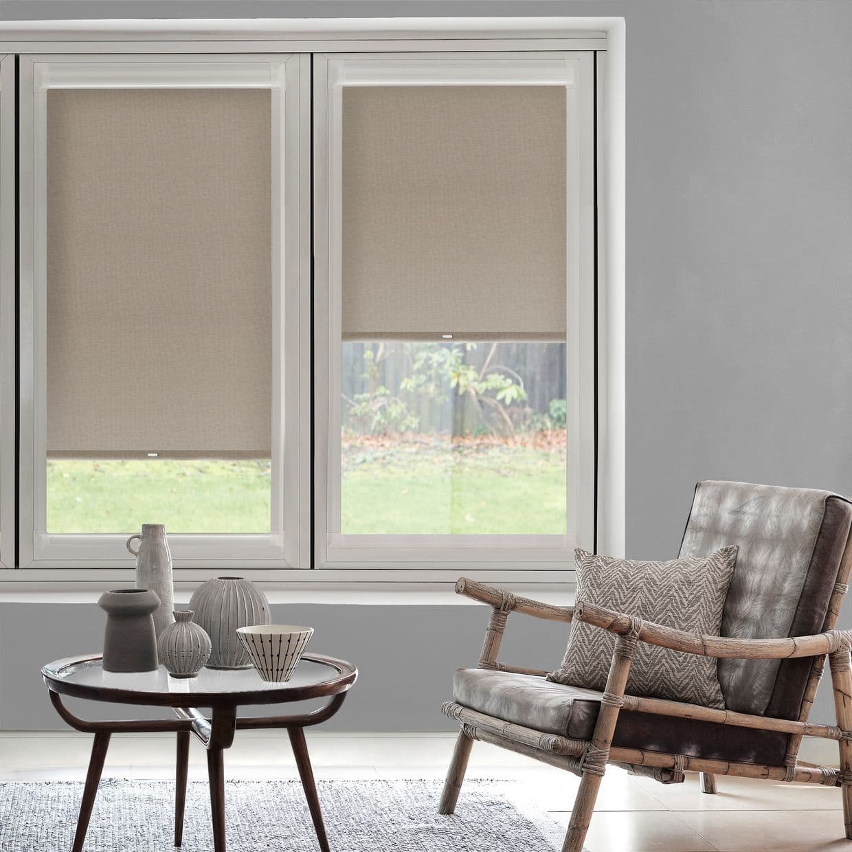Beige roller blinds in two adjacent windows, partially lowered and filtering daylight; above a sill with vases, beside a bamboo-frame leather armchair in a living room.