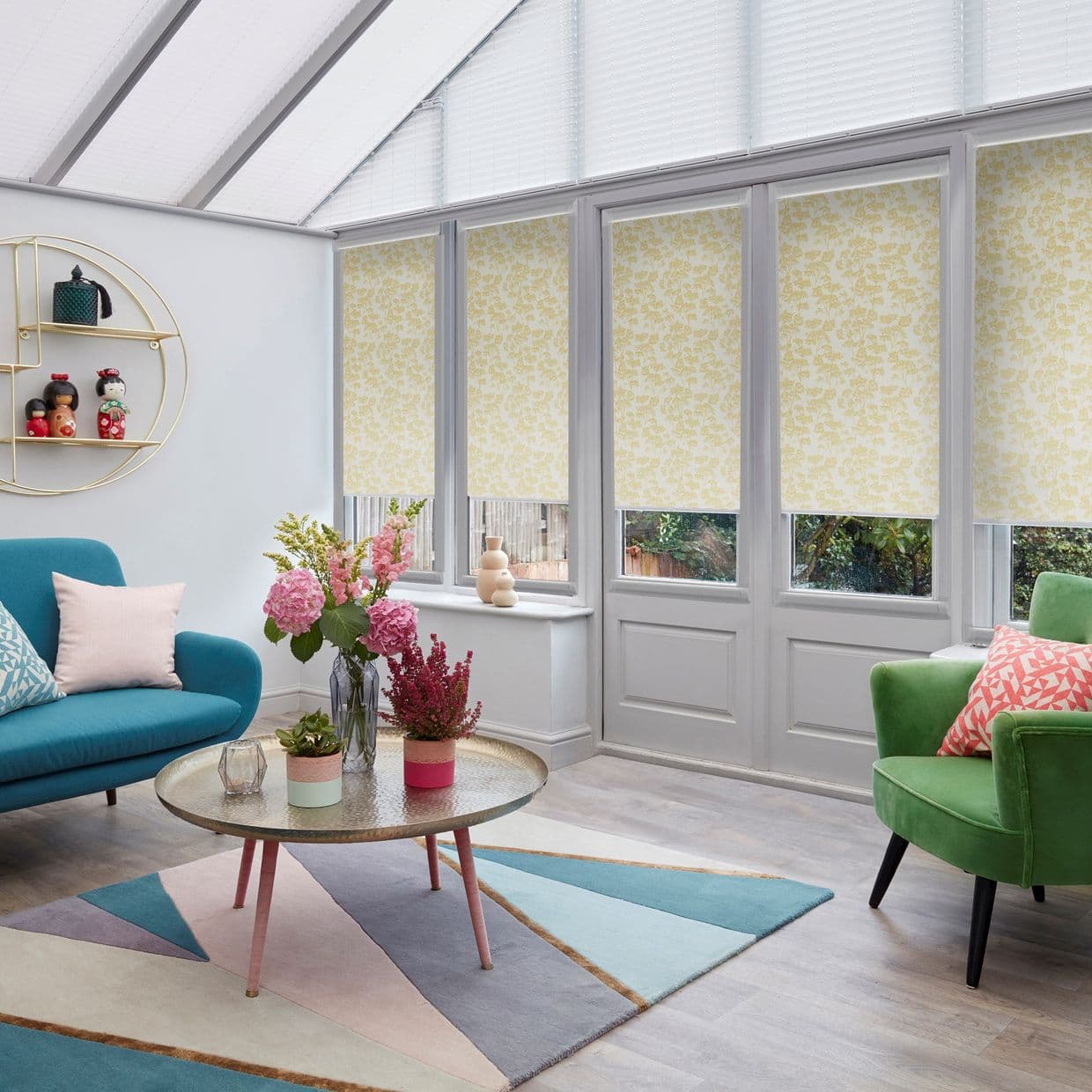Yellow floral roller blinds lowered across tall glazed doors and windows, filtering daylight, in a bright sunroom with roof glazing, teal sofa, green armchair, coffee table and geometric rug.