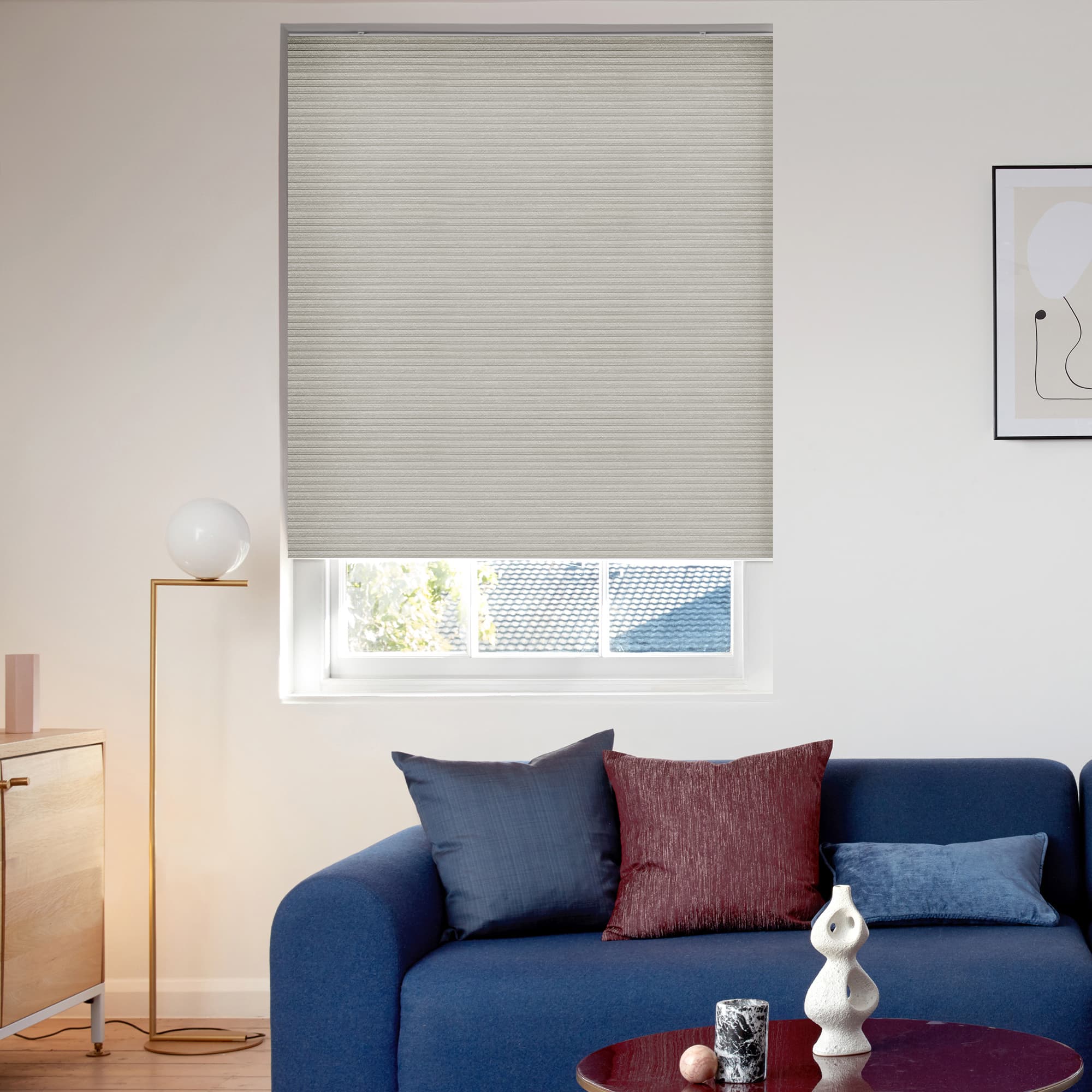 Beige cellular blind, lowered to cover the window with horizontal texture, filtering daylight; modern living room with blue sofa and cushions, brass floor lamp, small coffee table, framed wall art.