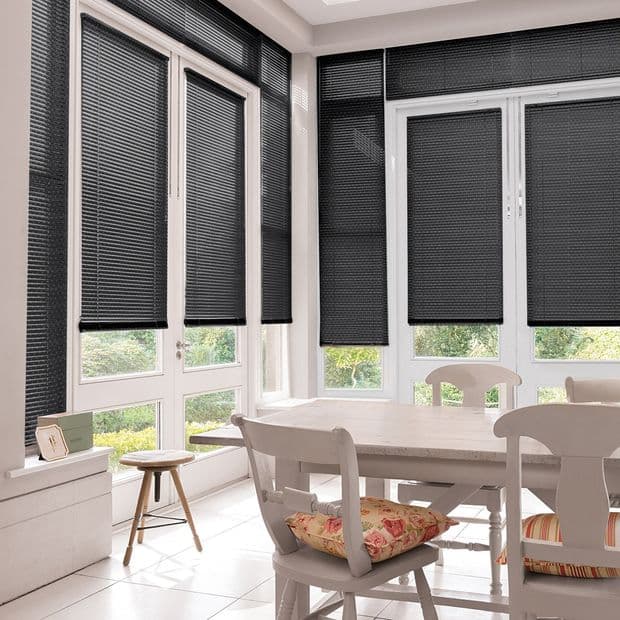 Black pleated blinds covering tall French windows and sidelights, lowered to varying heights, mostly closed but slightly raised at bottoms, filtering daylight in a bright dining area overlooking a garden.