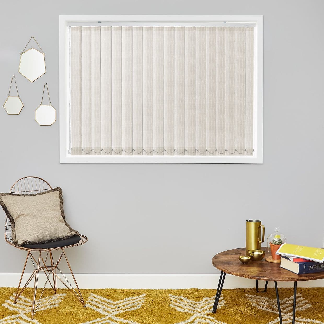 Beige vertical fabric blinds cover window, closed and softly filtering light in a white frame; grey living room wall with wire chair and cushion left, round wooden coffee table right.