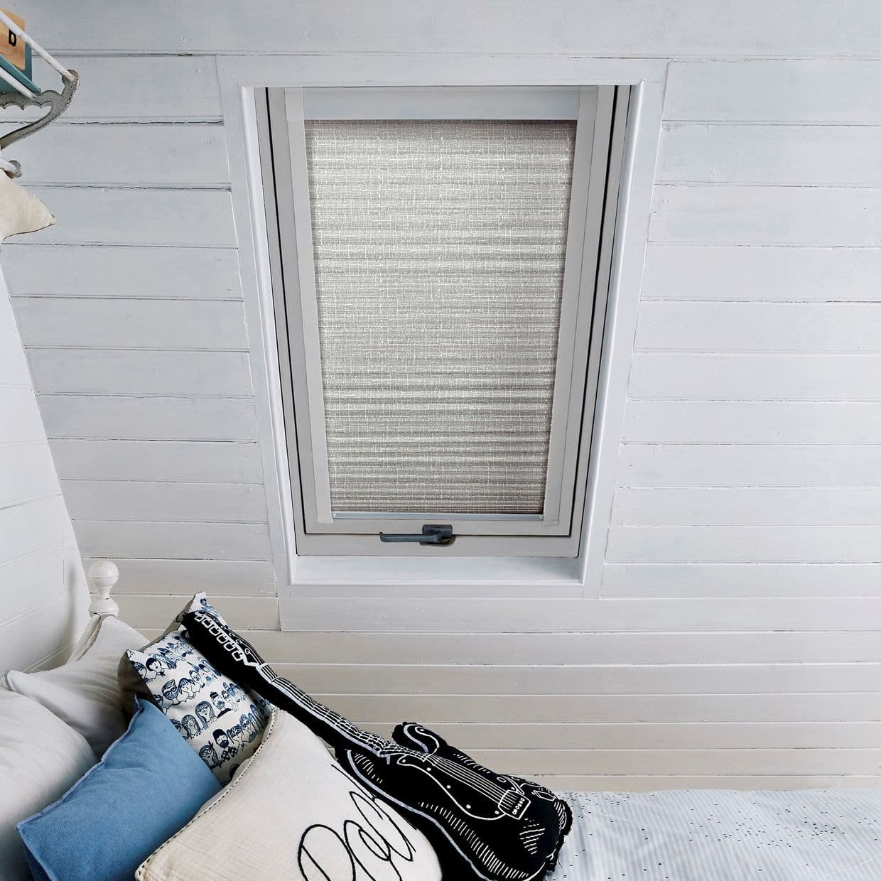 A pleated fabric blind on a skylight, fully lowered and filtering soft daylight; set in a white-painted slatted attic ceiling above a bed with pillows and a black guitar-shaped cushion.
