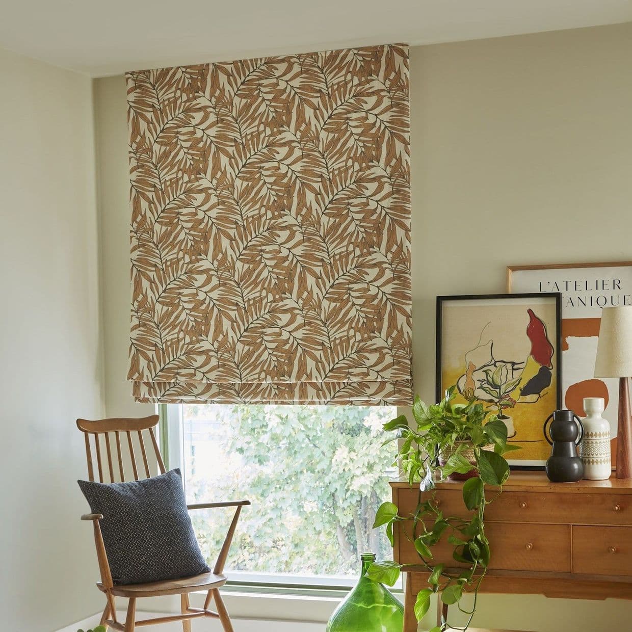 Roman fabric Roman shade with brown-and-cream leaf pattern, lowered and softly filtering daylight. Context: cozy living-room nook with wooden chair, cushion, sideboard, plants, and framed art.

L'ATELIER BOTANIQUE
