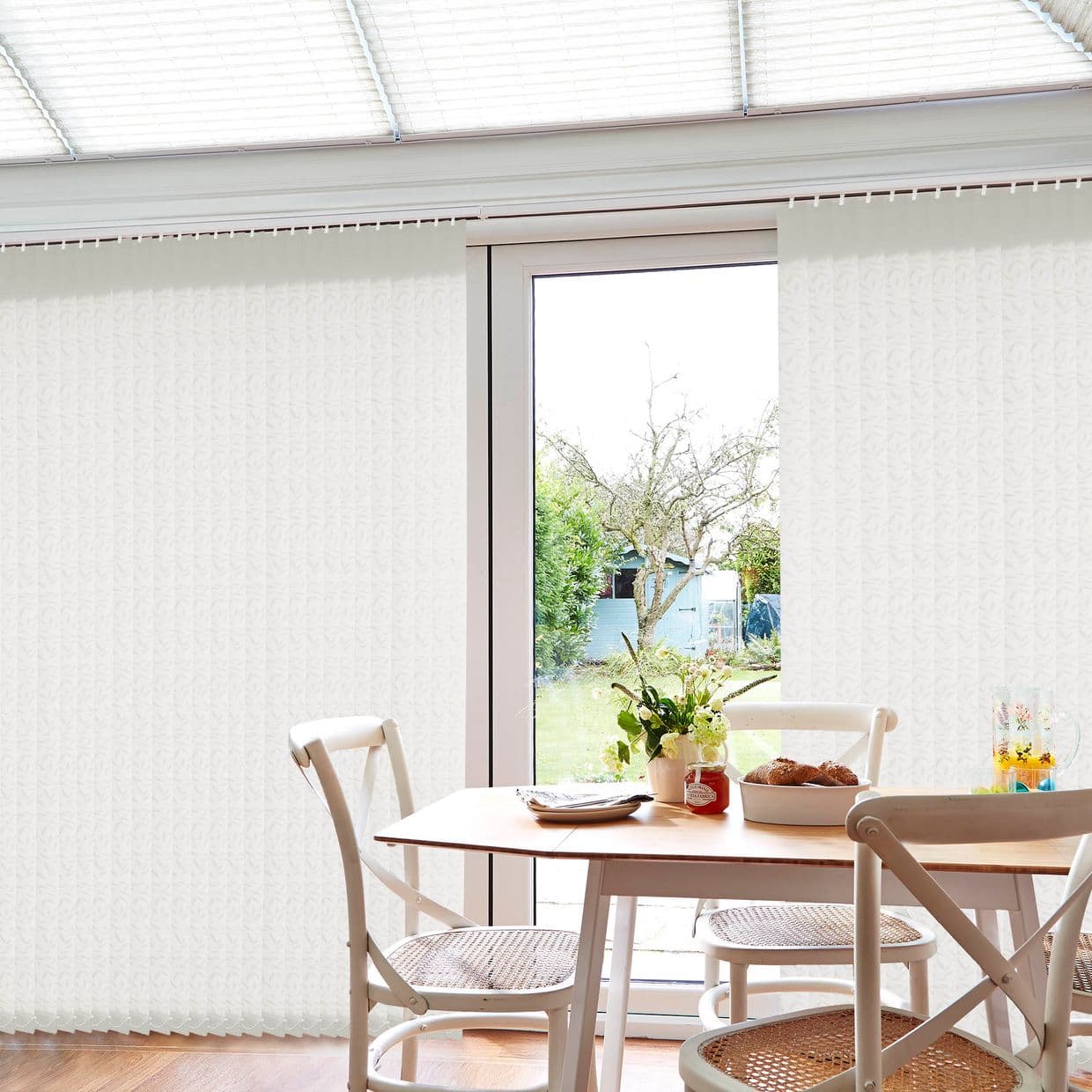 White textured vertical blinds, mostly closed but parted centrally to reveal a glass patio door; filtering bright daylight into a dining nook with wooden table, chairs, and backyard tree view.