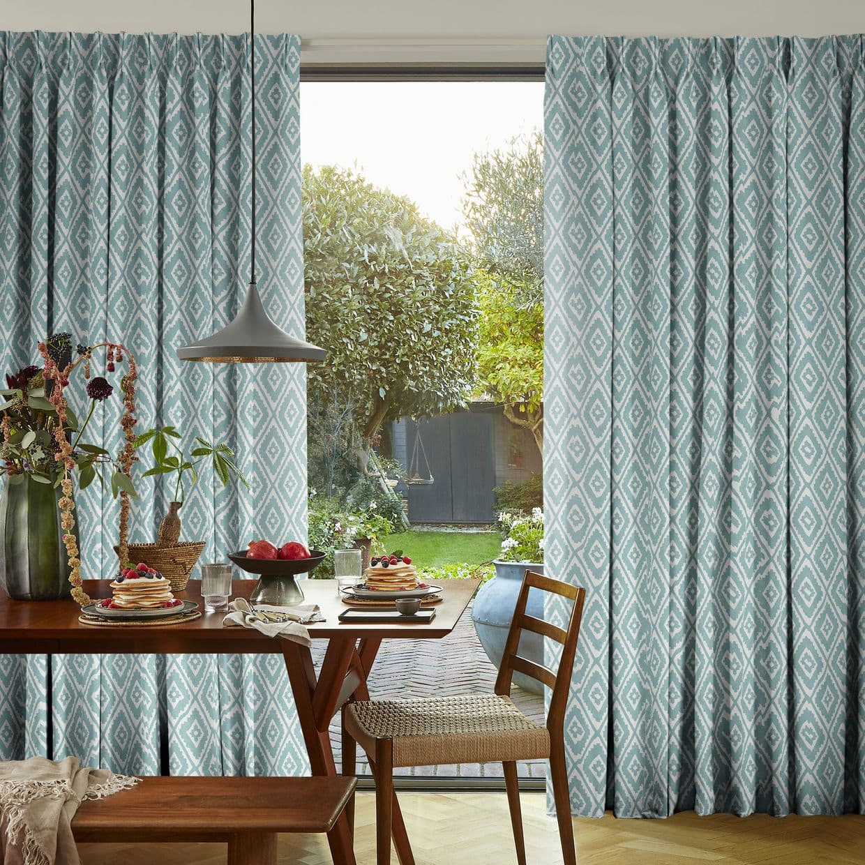 Teal-blue diamond-patterned pleated curtains, hung full-length and parted to frame a sliding door; they expose a sunlit backyard. Dining table with wooden chairs, pendant lamp and breakfast setting foreground.