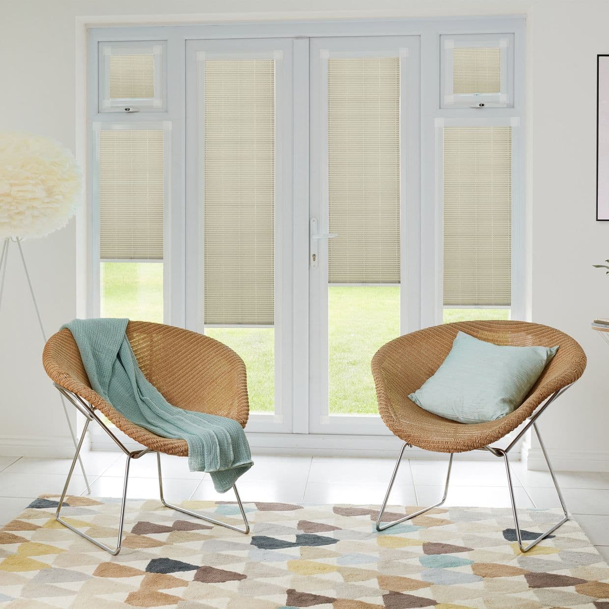 Beige pleated blinds lowered over French doors and sidelights, filtering daylight; two wicker chairs with teal throw and cushion on patterned rug in bright sitting room overlooking a lawn.