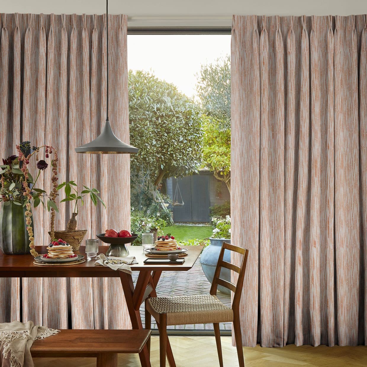 Patterned pleated drapery curtains, drawn partly aside revealing a central opening, softly filtering daylight; dining table with breakfast setting, pendant lamp and wooden chairs in a bright room overlooking a leafy garden.