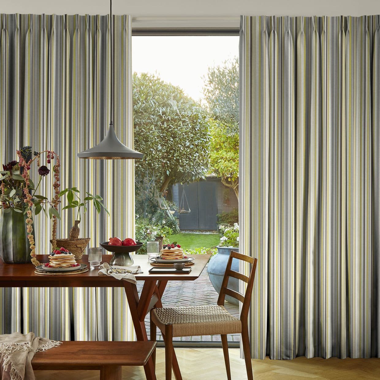 Floor-to-ceiling pleated striped curtains (gray, white, yellow) frame a central glass door, parted to reveal sunlight and a leafy backyard; foreground dining table set with pancakes, pendant lamp, wooden chairs.