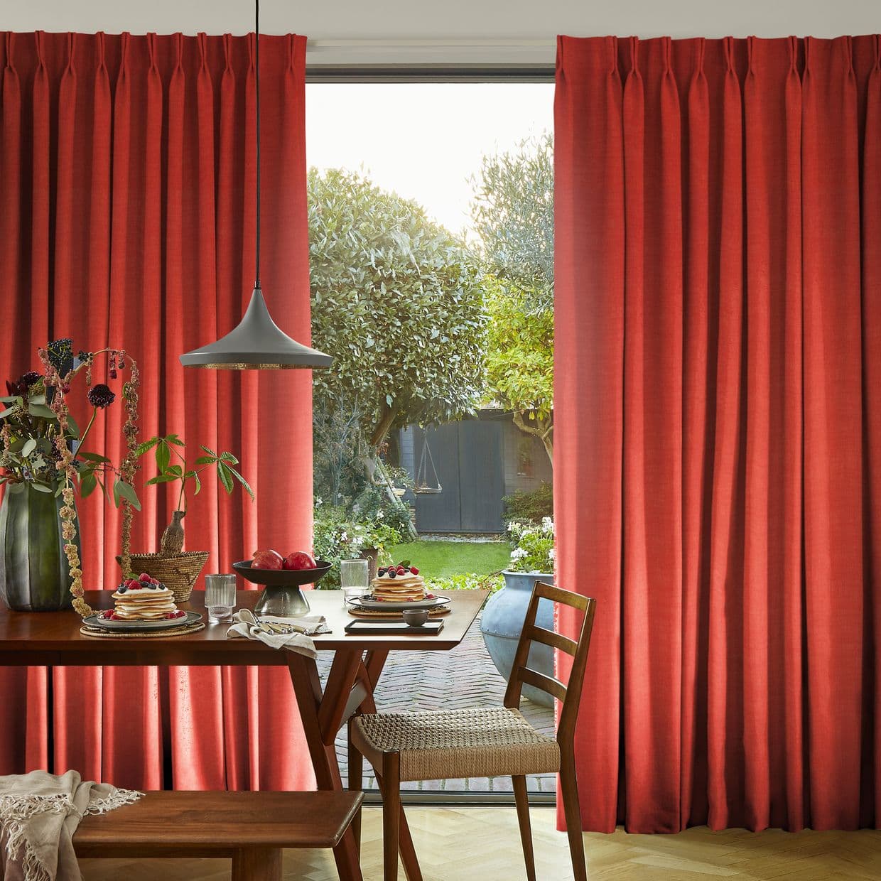 Red pleated floor-length curtains, slightly parted, framing a glass door and admitting daylight and a leafy garden; a wooden dining table with pancakes, pendant lamp and chairs in foreground.