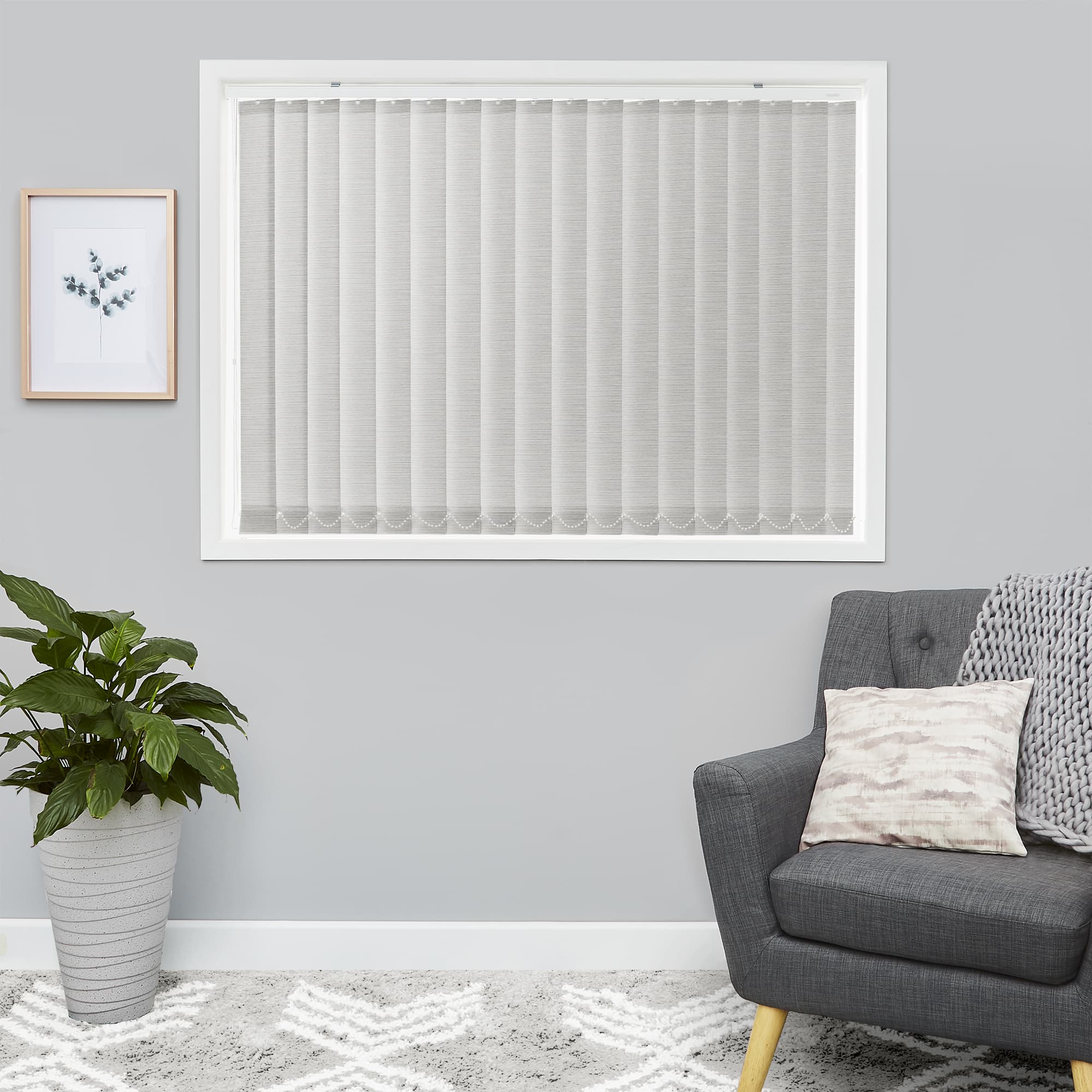 Light-gray vertical fabric blinds covering a window, closed and filtering daylight; set in a minimalist gray living room with a dark-gray armchair, rug, potted plant, framed botanical print.