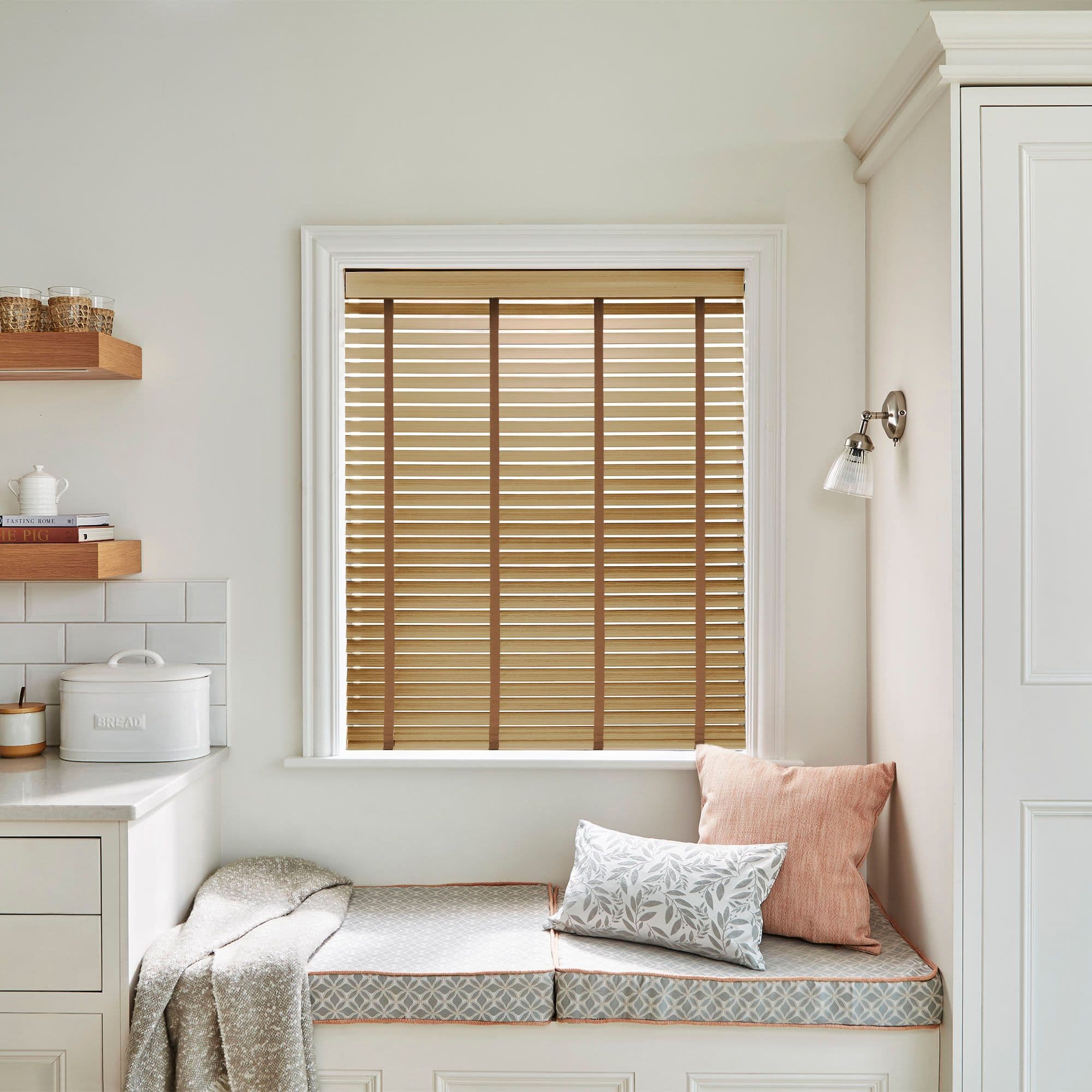 Horizontal wooden slatted blinds with brown ladder tapes â€” lowered and closed, filtering warm daylight â€” above a cushioned window seat in a pale, softly lit kitchen nook.

BREAD
THE PIG