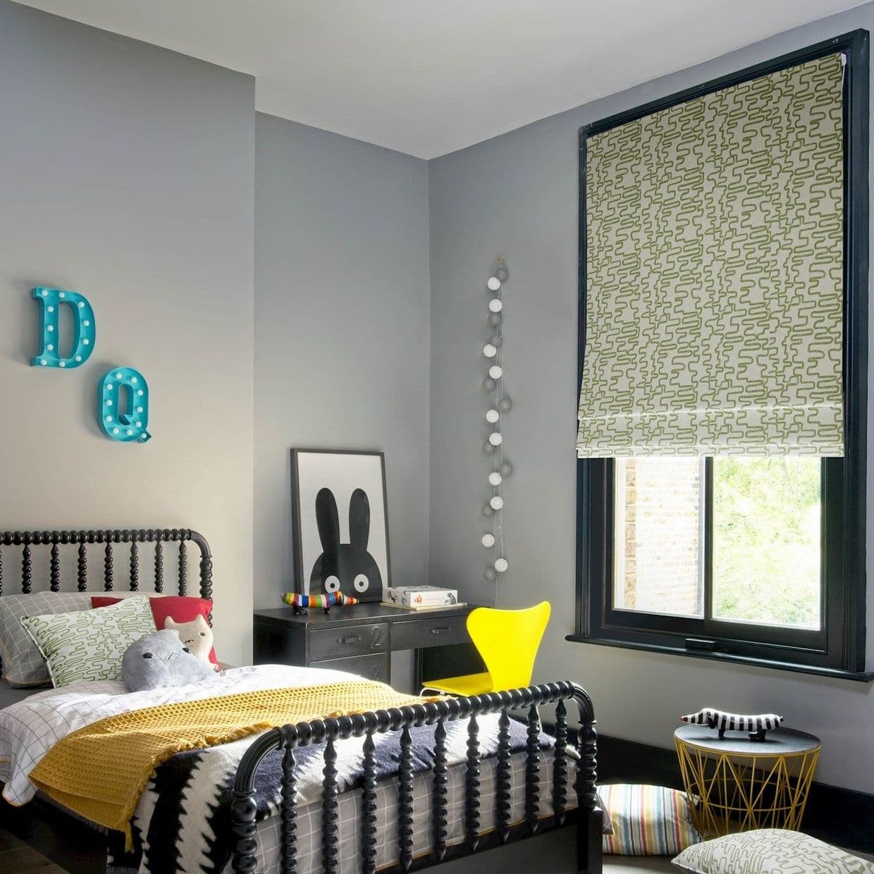 Green-and-white patterned Roman shade lowered over a black-framed window, filtering daylight; childâ€™s grey-painted bedroom with black spindle bed, yellow chair, desk, plush toys and string lights.