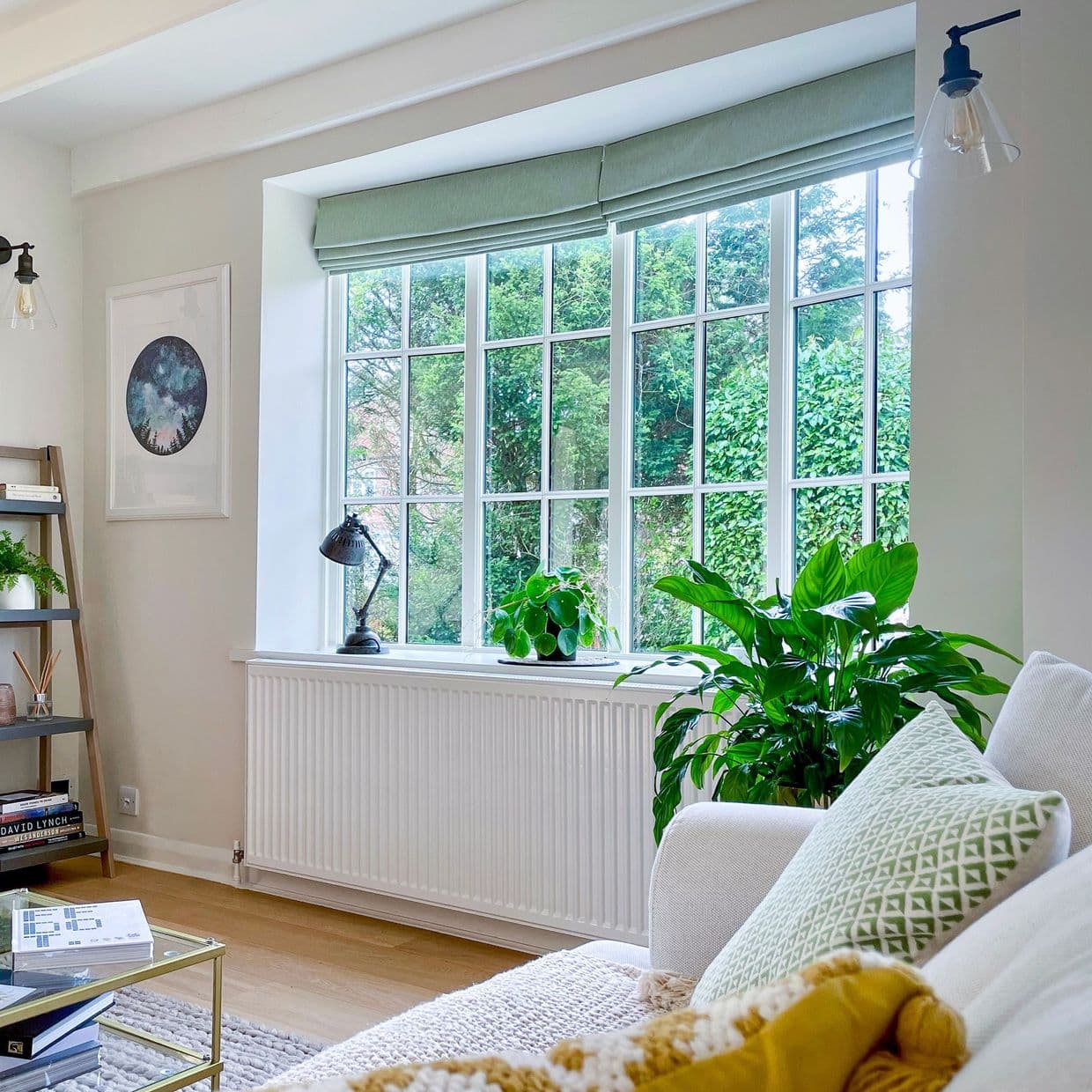 Green fabric Roman blind at window top, partially folded and filtering daylight; overlooks dense garden, with plants on the sill in a bright living room.