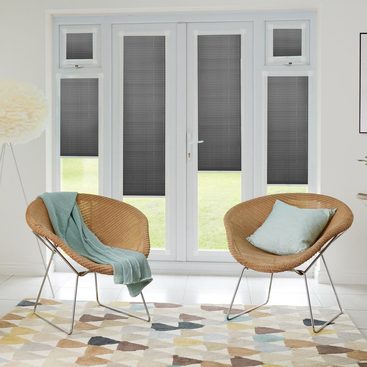 Dark gray pleated blinds installed inside French doors and side windows, lowered to block view and filter daylight; living room with two wicker chairs, pastel cushions, geometric rug, garden beyond.
