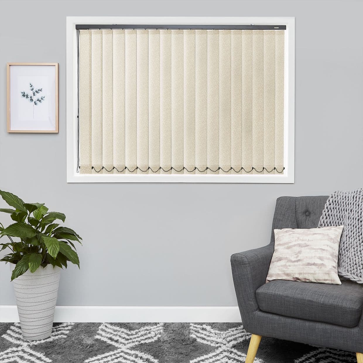 Vertical beige vertical fabric blinds cover the window, closed and gently filtering light with a beaded chain along the bottom, set in a gray-walled living room with plant, framed print, and armchair.