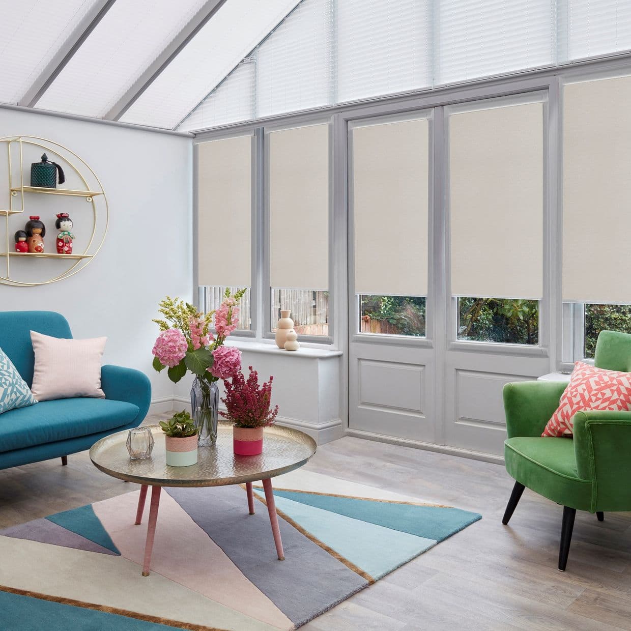 Beige roller blinds (four narrow panels) lowered over tall glazed doors and windows, filtering daylight; conservatory-style living room with teal sofa, green armchair, pastel geometric rug and round coffee table.