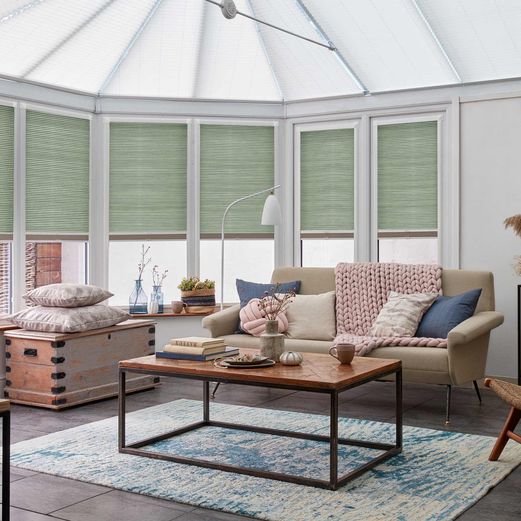 Pleated green cellular (honeycomb) blinds covering tall white-framed conservatory windows, lowered halfway and filtering soft daylight; surrounding light-filled living area with beige sofa, wooden coffee table, textured rug.