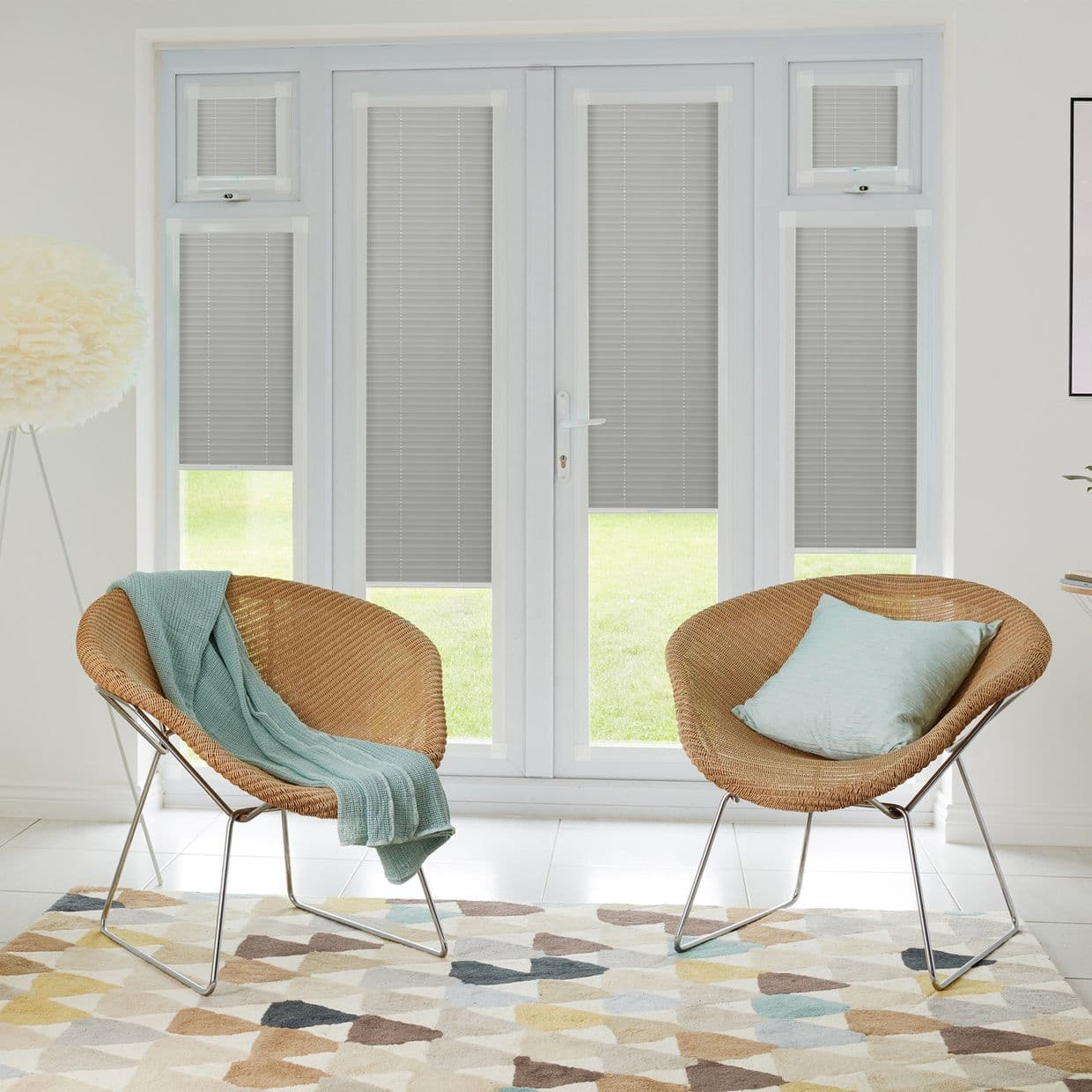 Grey pleated blinds inside white French doors and sidelights, lowered three-quarters, partially filtering light; bright sitting room with two wicker chairs, pastel cushions, patterned rug, lawn outside.