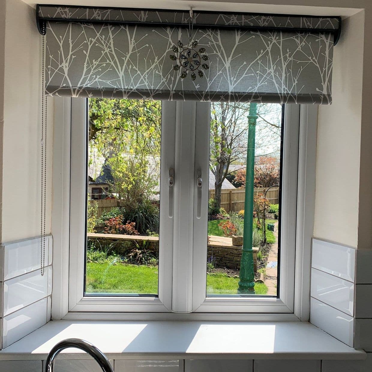 Patterned roller blind with white branch print hangs partially down over casement window, filtering light; kitchen sink and tiles frame a sunny backyard with lawn, plants and green lamp post.