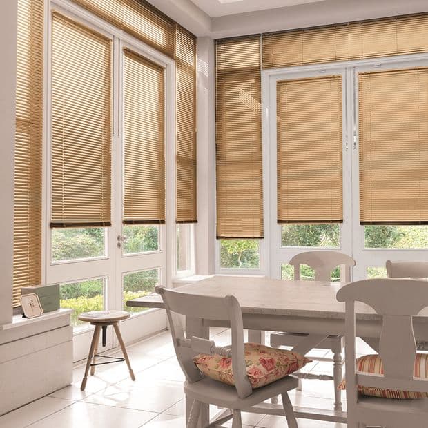 Tan horizontal Venetian blinds cover floor-to-ceiling windows and doors, lowered and filtering warm sunlight; bright dining sunroom with white wooden table and chairs, floral cushions, tiled floor and garden view.