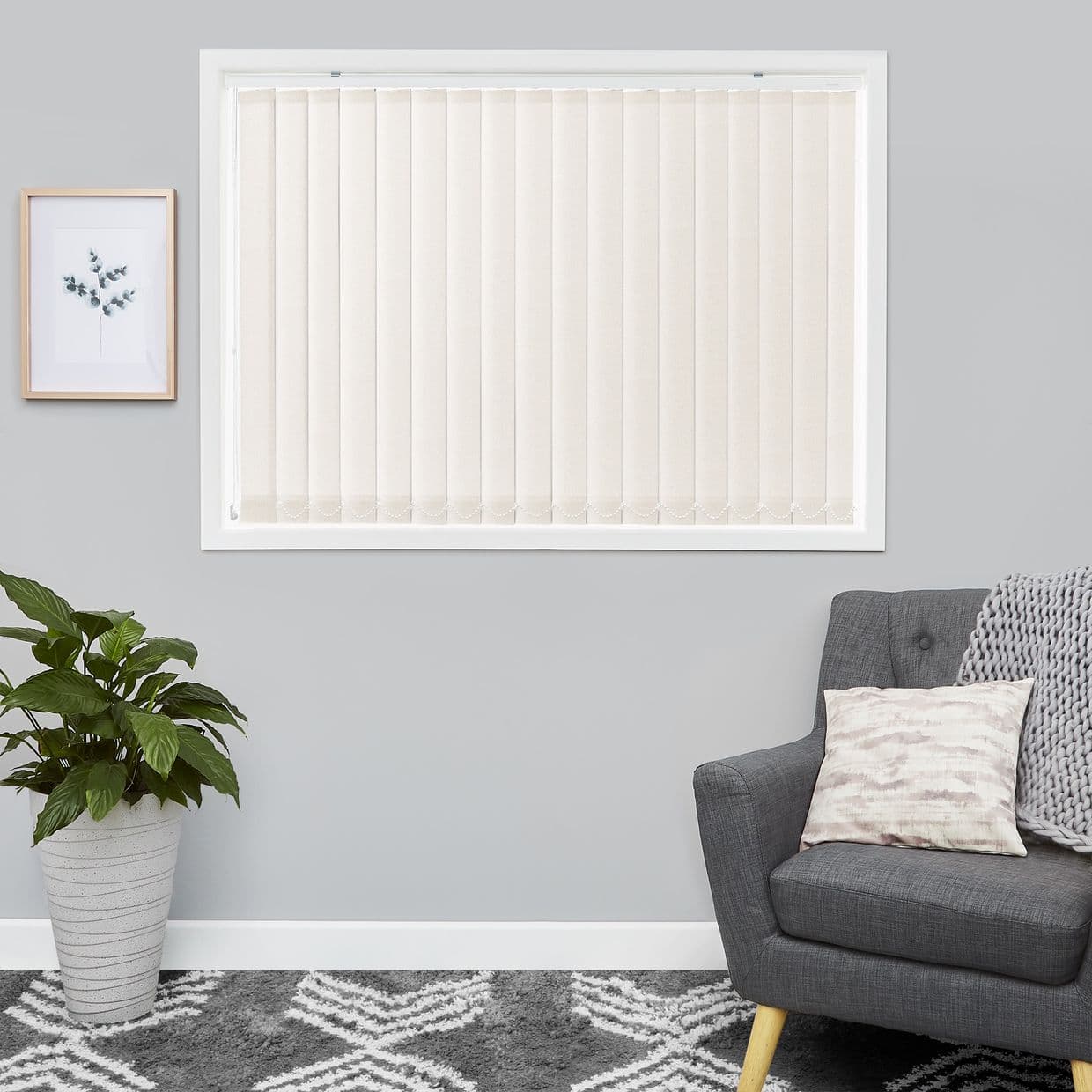 Cream vertical blindsâ€”fully closed, textured slatsâ€”cover a white-framed window, softly filtering daylight; set in a gray living room with potted plant, framed print, gray armchair.