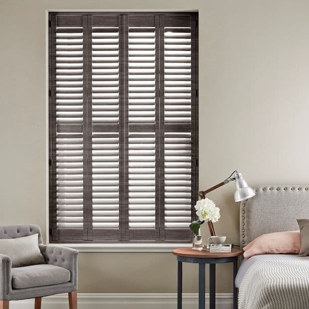 Floor-to-ceiling wooden plantation shutters covering the window, closed with angled louvers filtering daylight; set in a neutral bedroom with upholstered bed, nightstand, lamp, vase of flowers and armchair.