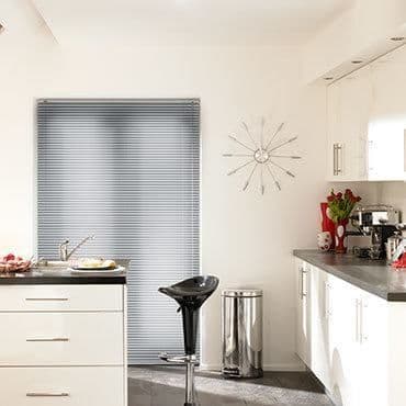 Metallic horizontal venetian blinds, fully lowered and closed, filtering soft daylight; modern white kitchen with black countertop, bar stool, stainless bin, wall clock, coffee machine, and red tulips.