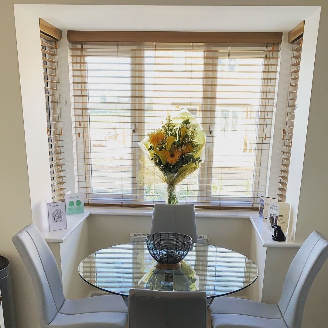 Wooden horizontal blinds across a bay window, slats tilted to filter bright daylight; small dining bay with round glass table, four grey chairs and a bouquet of yellow flowers on the sill.