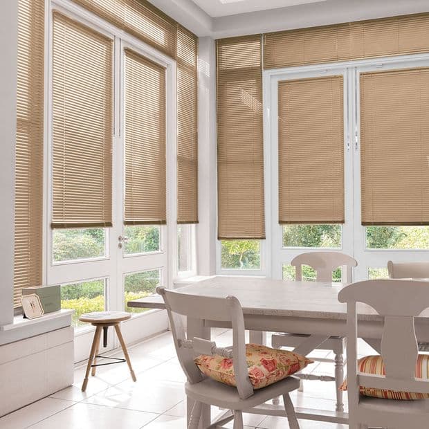 Beige horizontal blinds cover tall corner windows, slats mostly closed with low raised bottoms; filtering soft daylight into a bright dining nook with white table, chairs and floral cushions.
