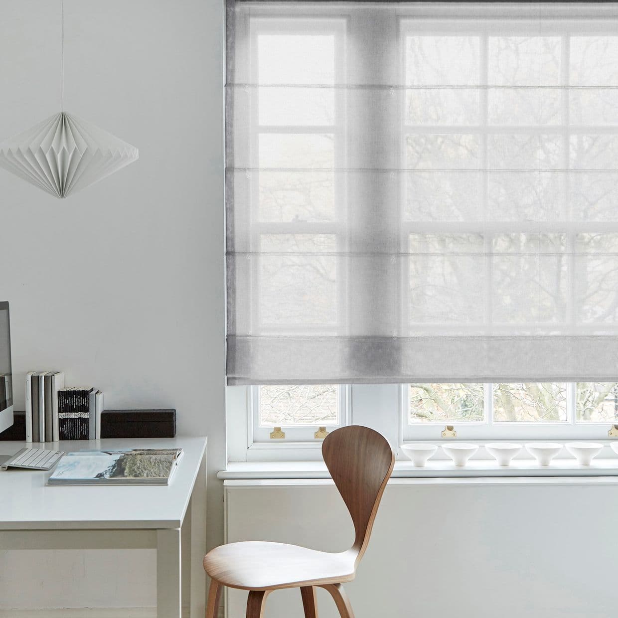 Sheer grey Roman blind lowered over a multiâ€‘pane window, softly diffusing daylight; white desk with books and wooden chair beneath, pendant lamp left, pale minimalist room with treed view.
