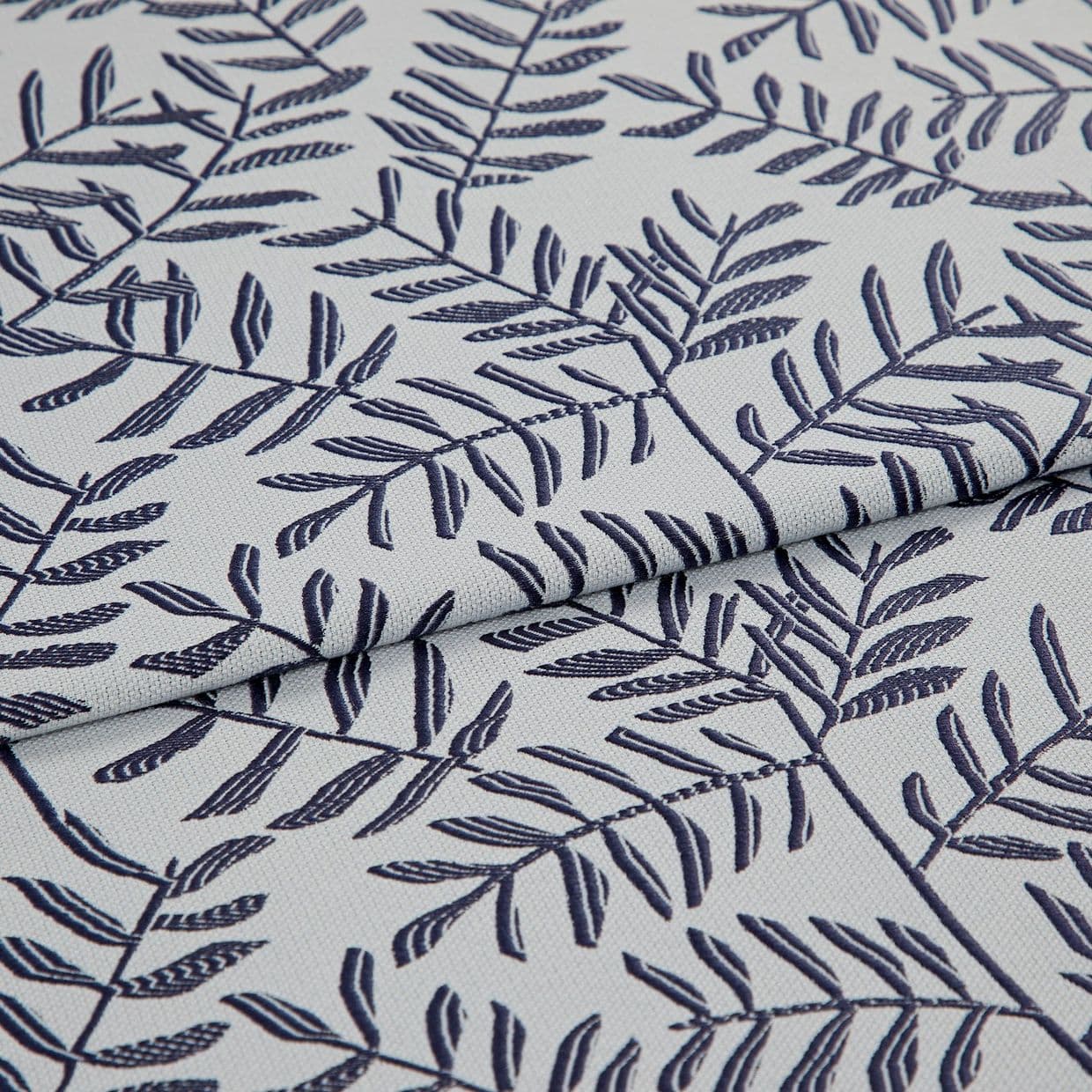 Light gray woven fabric with navy embroidered fern-like leaf pattern, folded in the center creating overlapping layers; close-up, soft diffuse lighting reveals textile texture and stitch detail.