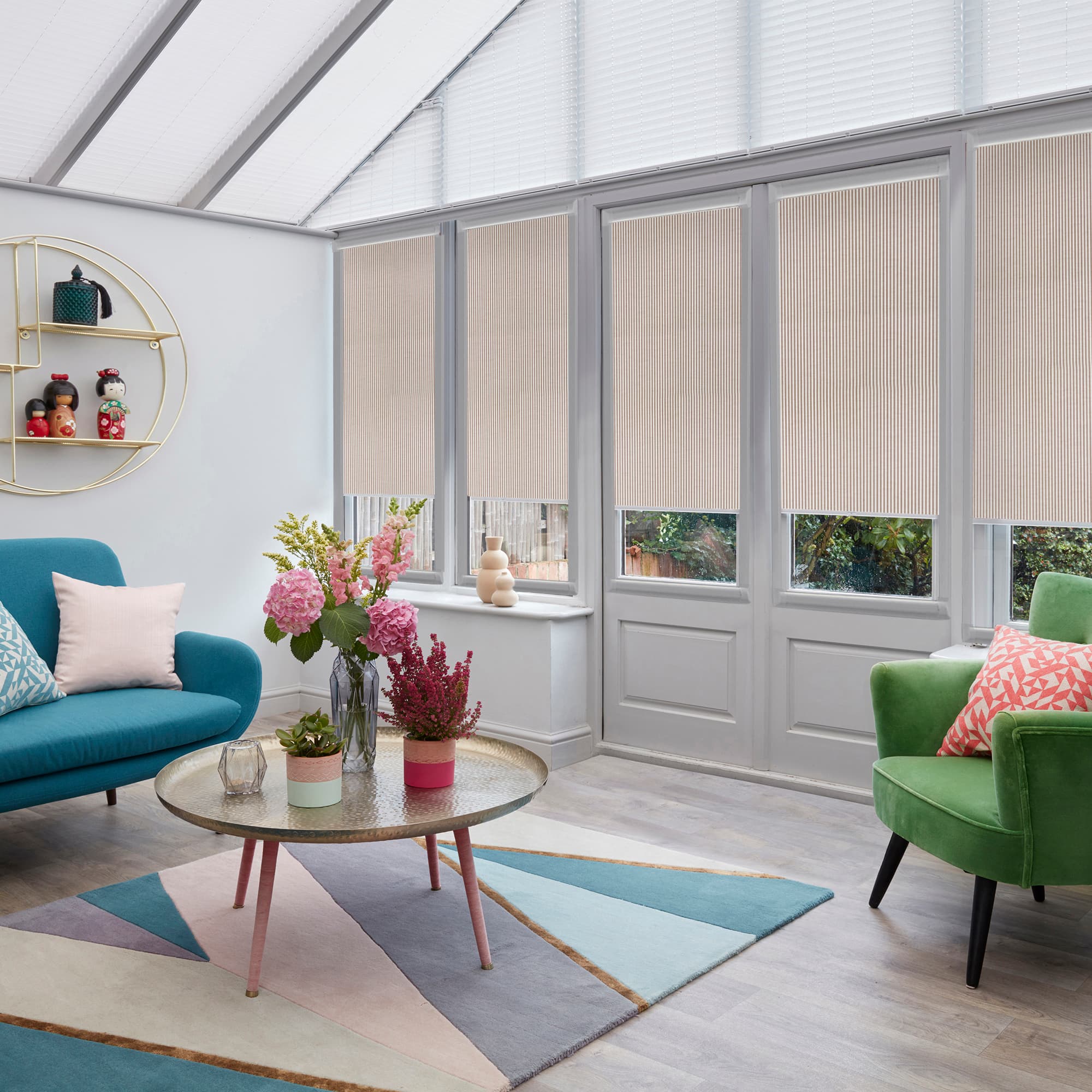 Beige pleated blinds with vertical ribbing, lowered over tall glass doors and windows to just above sills, filtering daylight; bright conservatory room with teal sofa, green armchair, pastel decor.