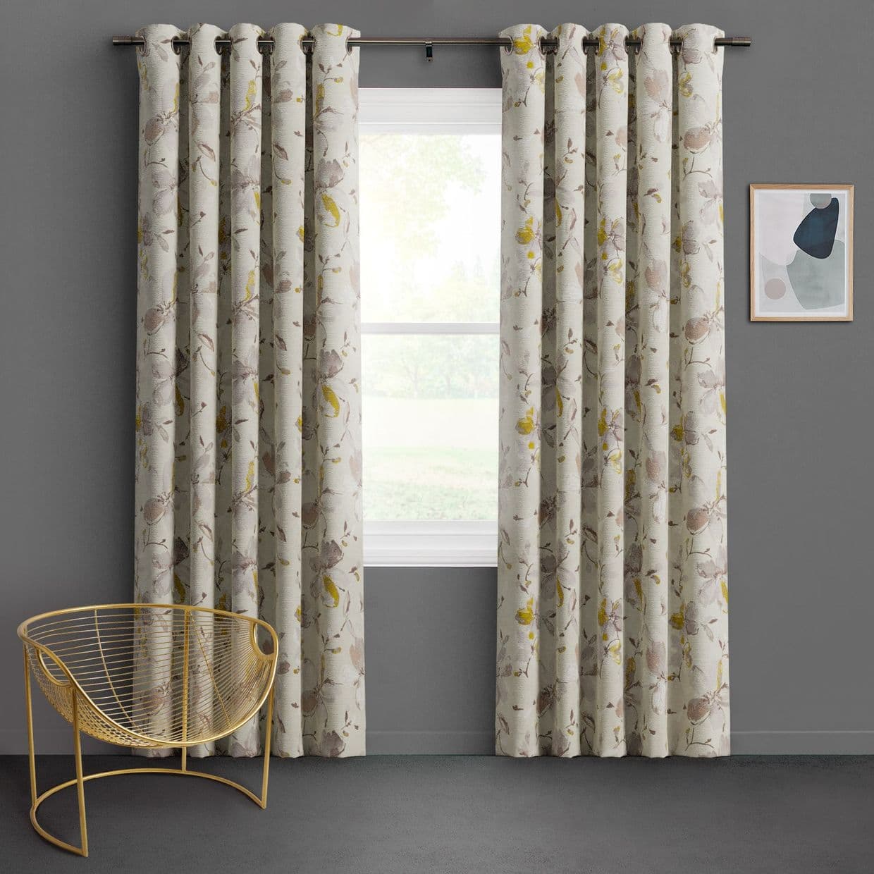 Grommet-top floral curtains, cream with muted beige and yellow blossoms, hang in pleats framing a central window, filtering soft daylight in a gray-walled room with a gold wire chair.