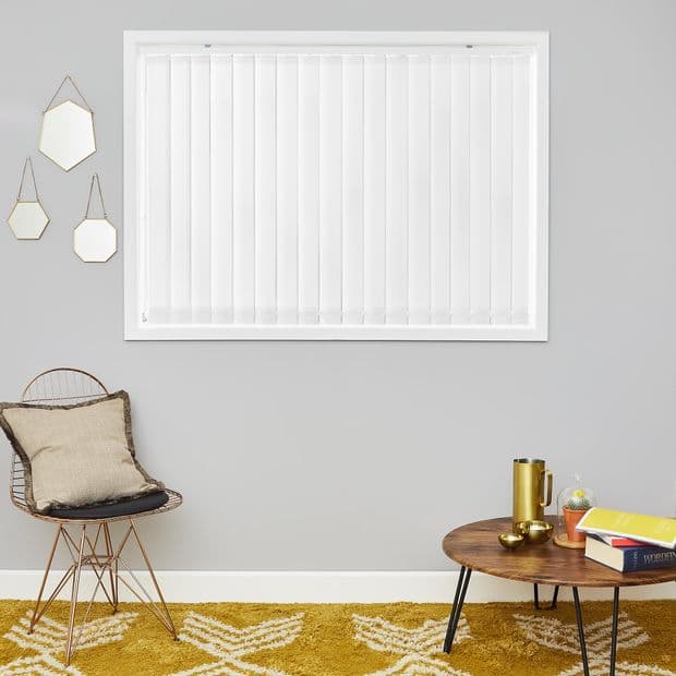 White vertical blinds covering a centered window, closed and aligned, softly diffusing daylight; neutral living room with gray walls, mustard rug, wireâ€‘frame chair with cushion, low wooden coffee table.