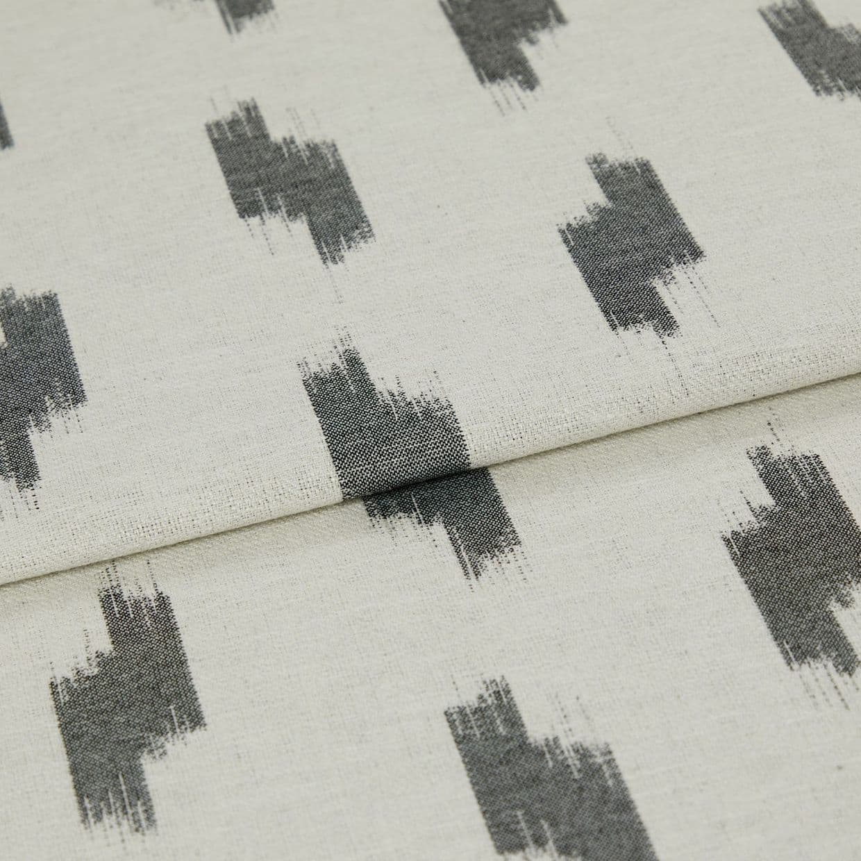 Folded curtain fabric (linen-like) with a central horizontal fold, showing repeating dark gray, brushstroke rectangular motifs on a light beige background in soft, even lighting.