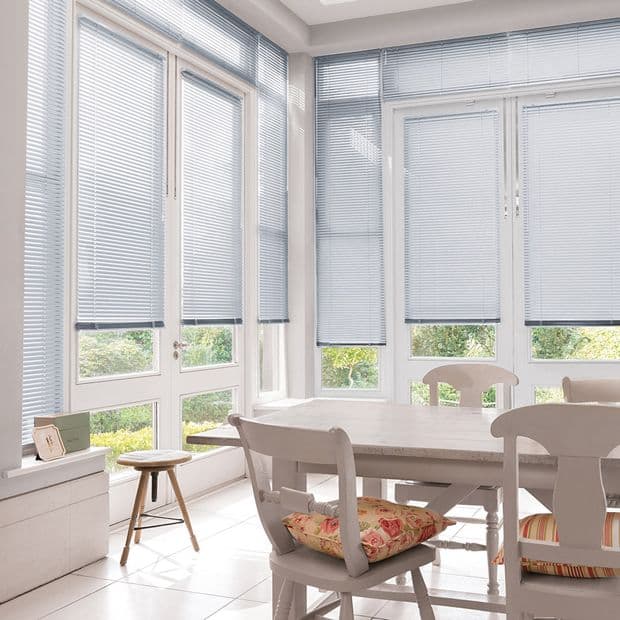 Horizontal light-gray Venetian blinds, on corner floor-to-ceiling windows and doors, slats partially closed; filtering daylight and revealing narrow garden views in a bright sunlit dining/sunroom with table and chairs.